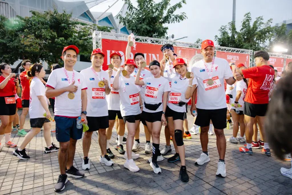 Group of runners in white shirts and red caps proudly displaying medals at a race finish, posing in front of a red backdrop.
